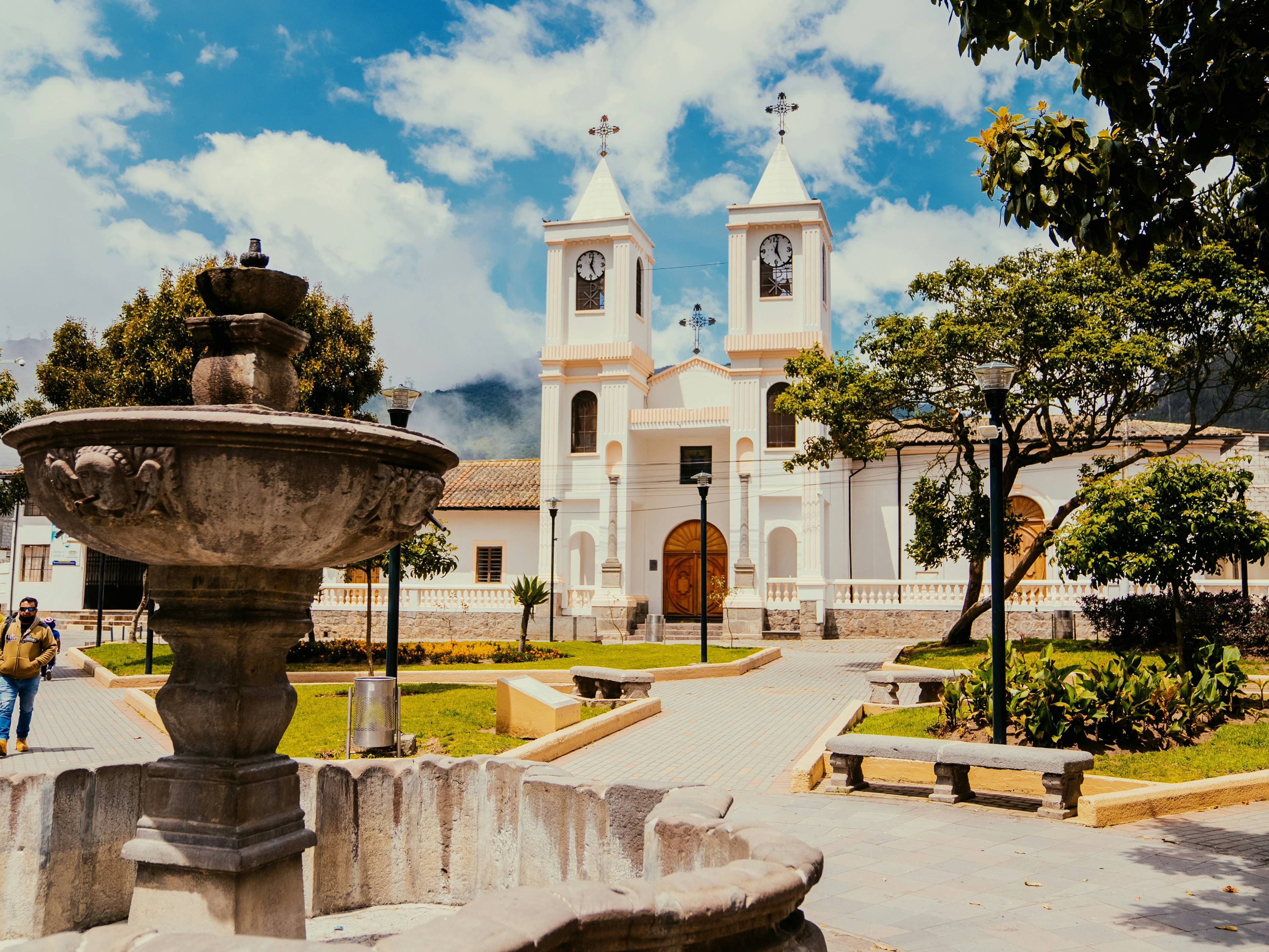 La imagen muestra una plaza con una fuente ornamental y una iglesia al fondo, rodeada de árboles y un cielo despejado.