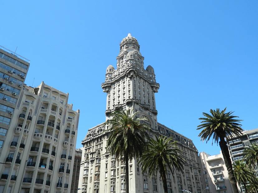 Vista de un imponente edificio en estilo arquitectónico ecléctico con detalles ornamentales, rodeado de palmeras y bajo un cielo despejado.