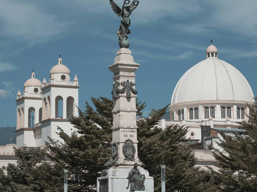Vista de un monumento con una estatua alada en primer plano y una cúpula de iglesia al fondo bajo un cielo despejado.