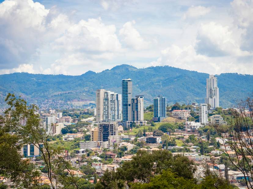 Vista panorámica de una ciudad moderna con rascacielos y un fondo de montañas bajo un cielo nublado.