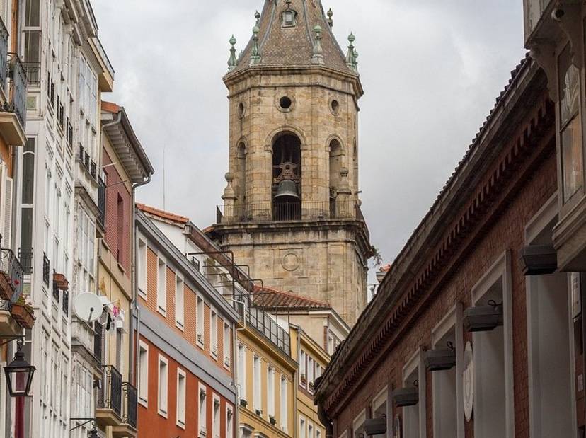 vitoria-3194558-1280-1_1 Una calle empedrada con edificaciones coloridas y una torre de iglesia al fondo bajo un cielo nublado.