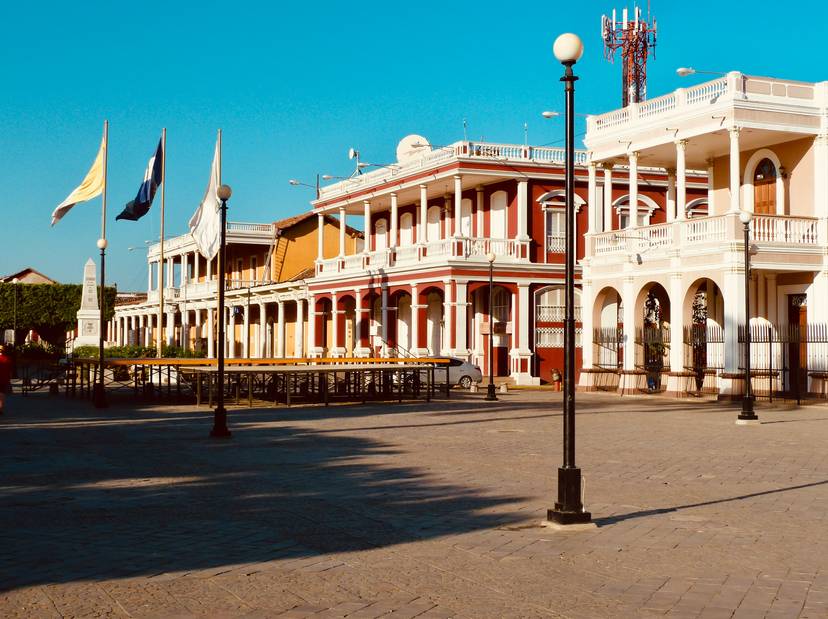Vista de una plaza pública con edificios de estilo colonial y banderas ondeando.