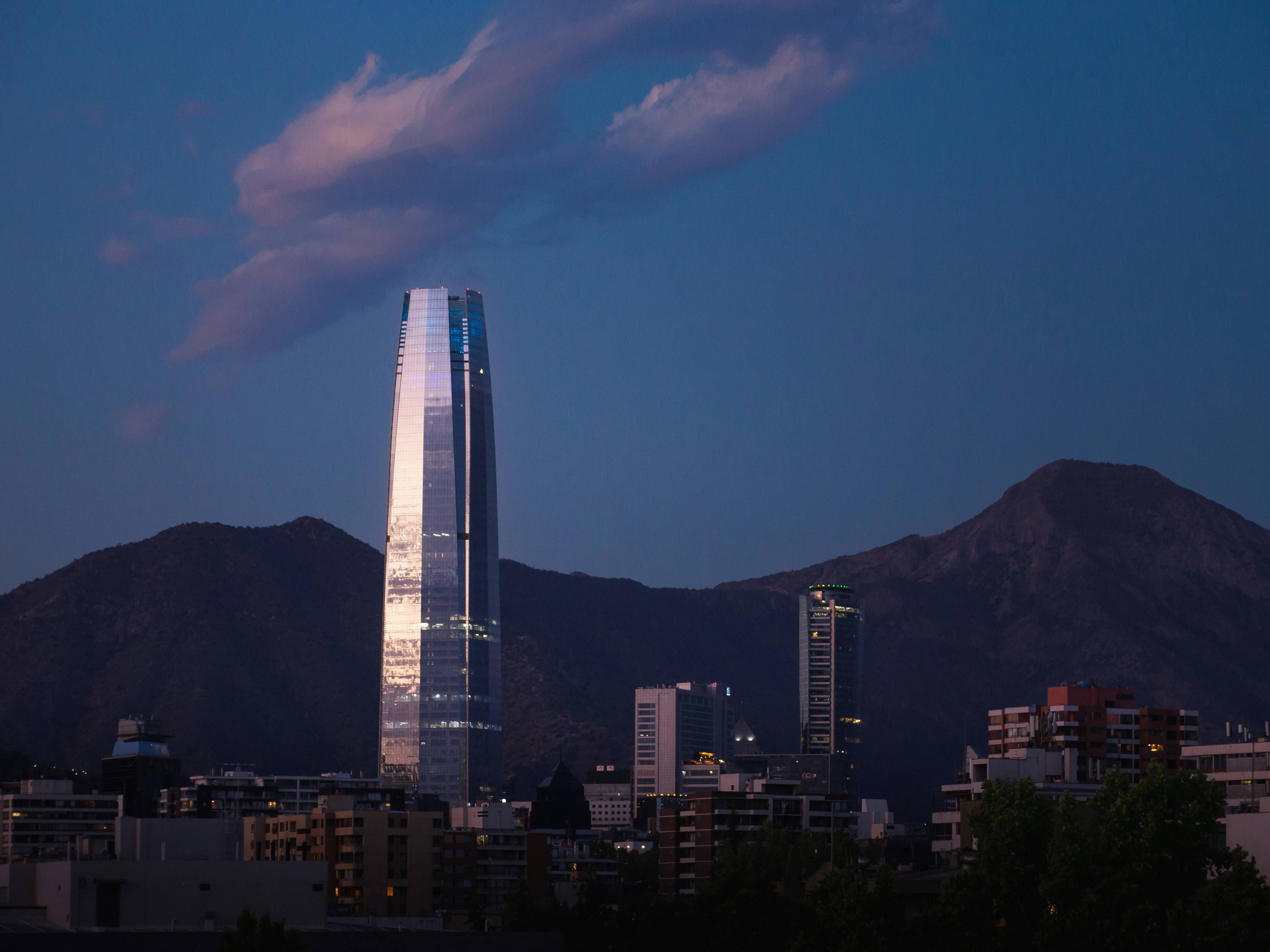 La imagen muestra un rascacielos moderno iluminado al atardecer en una ciudad rodeada de montañas.