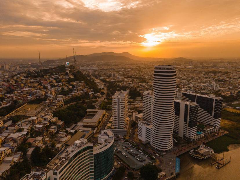 Vista aérea de una ciudad moderna al atardecer, destacando edificios altos y curvos con un fondo de montañas.