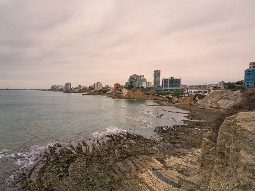 Vista costera de una ciudad moderna con edificios altos al fondo y rocas en primer plano bajo un cielo nublado.