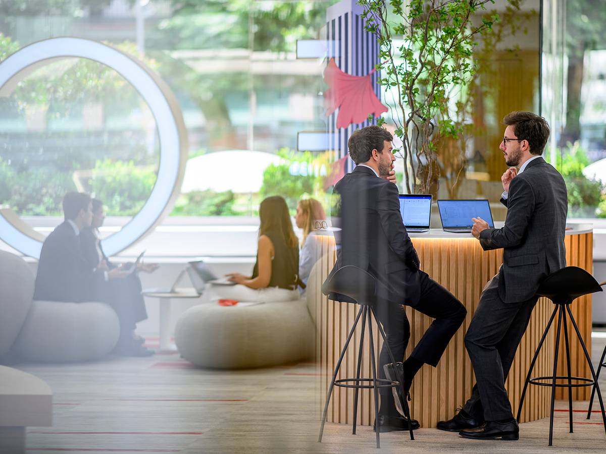 Talent Dos hombres conversan en un entorno moderno de oficina mientras otros trabajan en segundo plano.