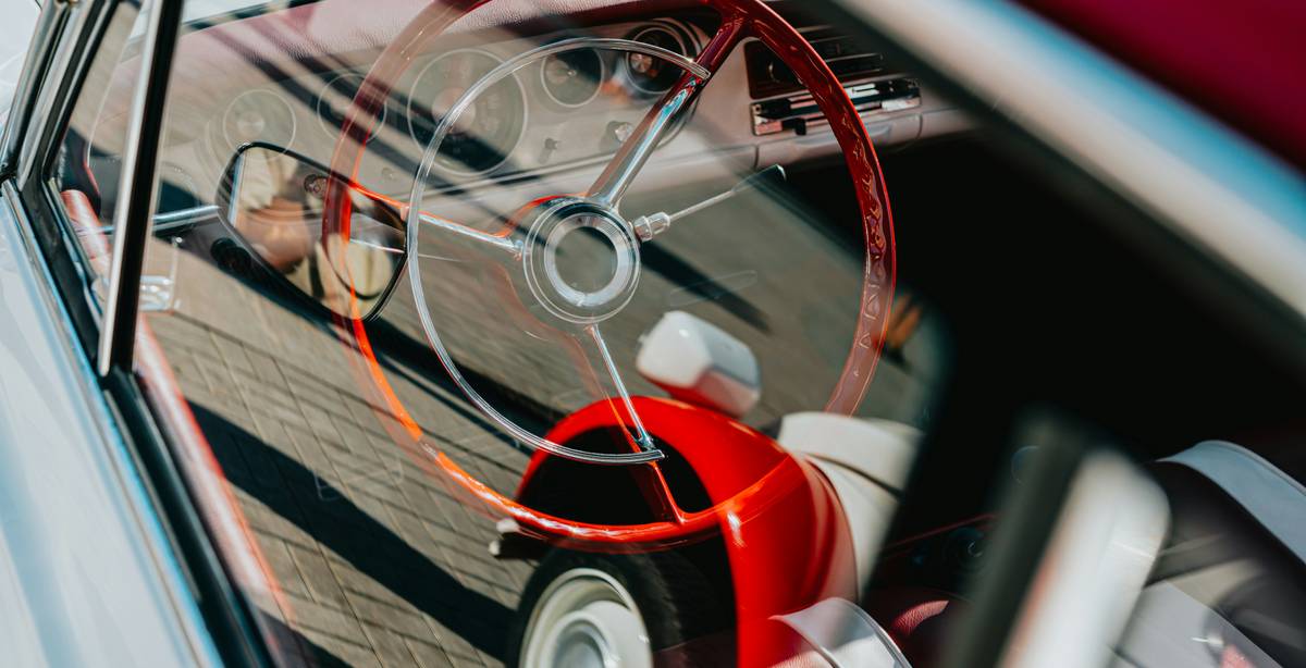 La imagen muestra el interior de un automóvil clásico con un volante de color rojo y asientos en tonos claros.