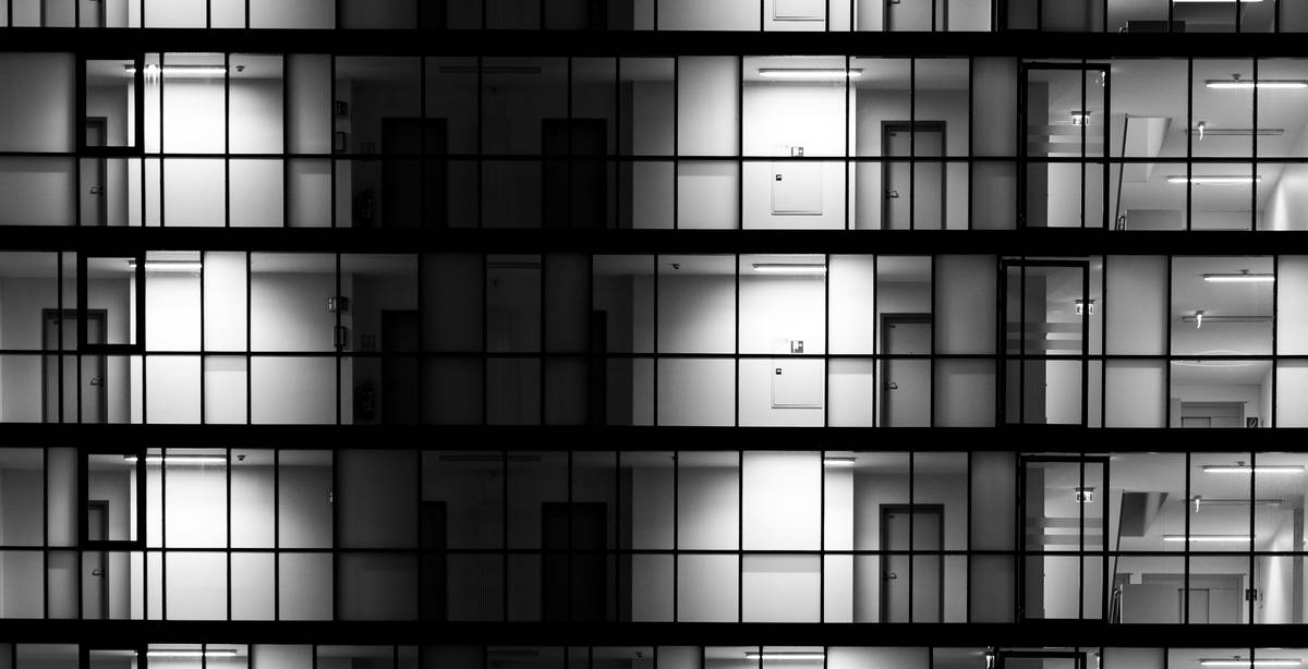 La imagen muestra una fachada de un edificio iluminado en blanco y negro, destacando filas de ventanas con distintos niveles de luz.