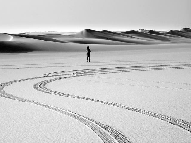 Una persona solitaria se encuentra en un vasto desierto, rodeada de onduladas dunas de arena bajo un cielo despejado.
